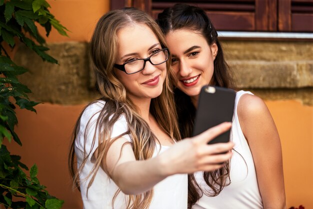 Deux belles filles de race blanche prennent selfie sur le smartphone près du mur d'un immeuble