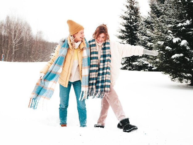 Deux belles jeunes femmes hipster souriantes dans des vêtements chauds à la mode et des écharpes. Femmes insouciantes posant dans la rue dans le parc. Des modèles purs positifs s'amusant dans la neige. Profiter des moments d'hiver. Notion de Noël