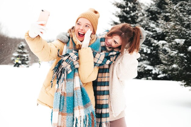 Deux belles jeunes femmes hipster souriantes dans des vêtements chauds à la mode et des écharpes. Femmes insouciantes posant dans la rue dans le parc. Des modèles purs positifs s'amusant dans la neige. Profiter des moments d'hiver. Prendre un selfie