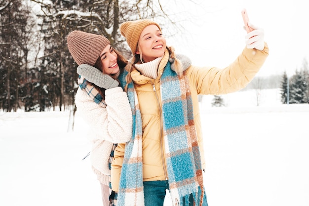 Deux belles jeunes femmes hipster souriantes dans des vêtements chauds à la mode et des écharpes. Femmes insouciantes posant dans la rue dans le parc. Des modèles purs positifs s'amusant dans la neige. Profiter des moments d'hiver. Prendre un selfie