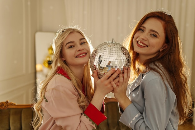 Deux belles jeunes filles caucasiennes en pyjama souriantes regardant la caméra tenant une boule disco devant elles Style de vie différentes émotions concept de loisirs