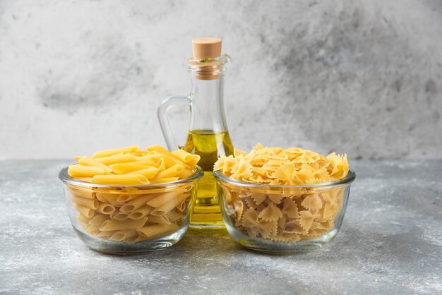 Deux bols de pâtes crues penne et farfalle avec une bouteille d'huile d'olive sur une surface en marbre.