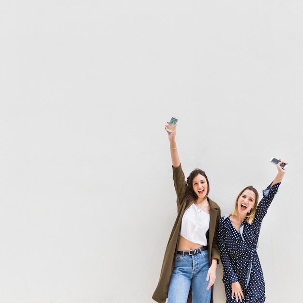 Deux élégante femme excitée, levant leur main tenant le téléphone portable sur fond blanc