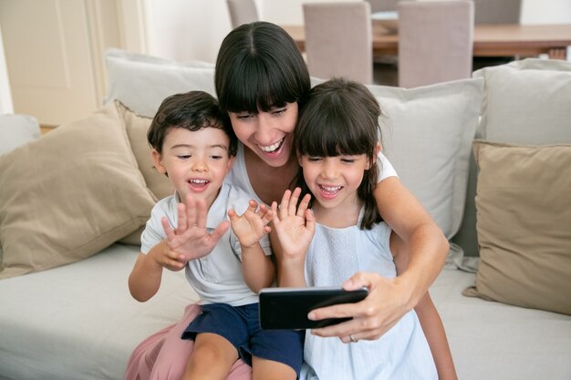 Deux enfants joyeux et leur maman heureuse à l'aide de téléphone pour un appel vidéo assis sur un canapé à la maison ensemble