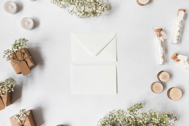 Photo gratuite deux enveloppes entourées de fleurs d'haleine de bébé; bougies; éprouvettes de guimauve; souches d'arbres miniatures et coffrets cadeaux sur fond blanc