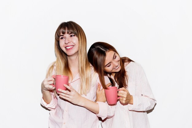 Deux femmes blanches joyeuses en pyjama rose avec une tasse de thé posant. Portrait flash.