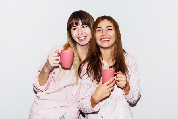 Deux femmes blanches joyeuses en pyjama rose avec une tasse de thé posant. Portrait flash.