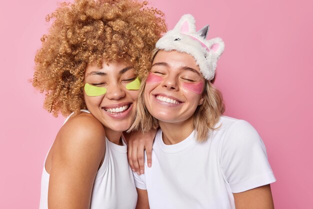 Deux femmes heureuses et amicales se tiennent étroitement l'une à l'autre appliquent des patchs de beauté sous les yeux subissent des procédures de soins de la peau à la maison gardent les yeux fermés sourient à pleines dents vêtues de vêtements décontractés posent à l'intérieur