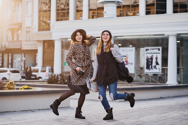 Deux femmes souriantes joyeuses à la mode sautant par-dessus la ville. Look élégant, voyager ensemble, porter des vêtements tendance modernes, marcher avec un café à emporter, exprimer des émotions positives.