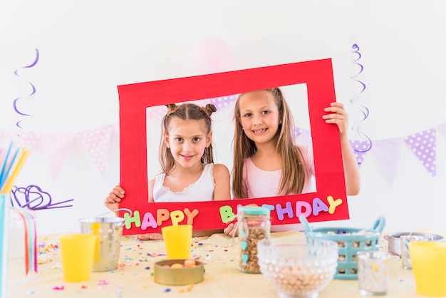 Deux fille souriante tenant cadre photo joyeux anniversaire avec des aliments différents sur la table