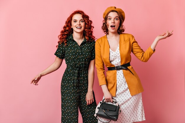 Deux filles excitées faisant des gestes tout en regardant la caméra. Curly femme française posant avec un ami sur fond rose.