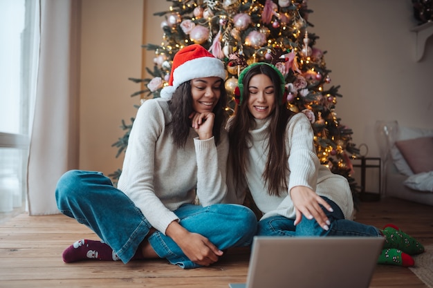 Deux filles souriant tout en parlant avec un ami en ligne sur un ordinateur portable pendant la célébration de Noël à la maison