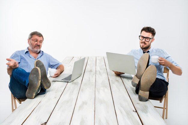 Les deux hommes d'affaires souriants avec les jambes sur la table travaillant sur des ordinateurs portables sur fond blanc. Des affaires à l'américaine