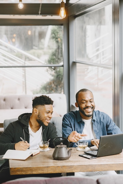 Deux hommes afro-américains travaillant derrière un ordinateur portable et écrivant dans un cahier. Hommes avec barbe assis dans un café.