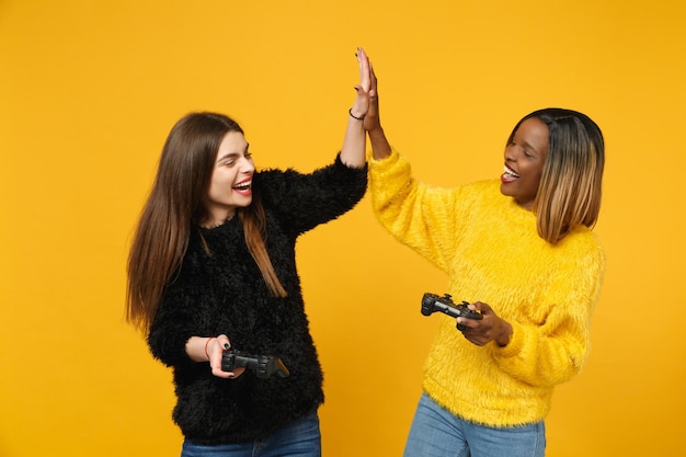 Deux jeunes amies amusantes européennes et afro-américaines en vêtements jaunes noirs tiennent le joystick isolé sur fond de mur orange vif, portrait en studio. concept de style de vie des gens. maquette d'espace de copie