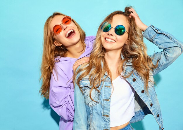 Deux jeunes belles blondes souriantes hipster femmes dans des vêtements d'été à la mode. Femmes sexy sans soucis posant près du mur bleu à lunettes de soleil. Modèles positifs