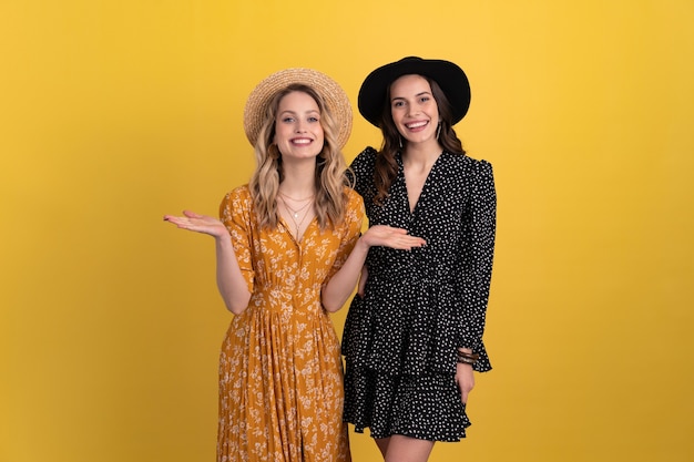 Deux jeunes belles femmes amies ensemble isolées sur fond jaune en robe noire et jaune et chapeau tendance bohème élégante, accessoires de style mode printemps été, souriante humeur joyeuse s'amusant