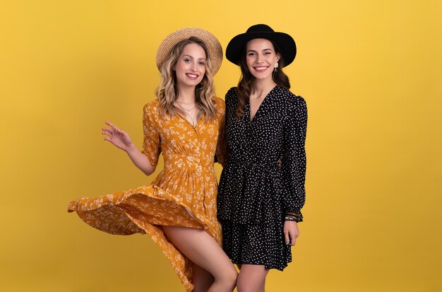 deux jeunes belles femmes amies ensemble isolées sur jaune en robe noire et jaune et chapeau tendance boho élégant