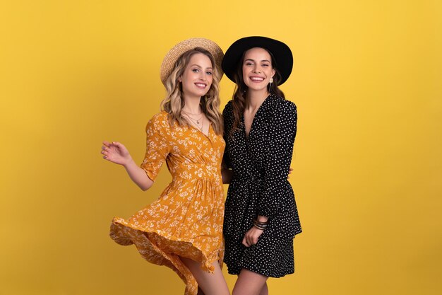 Deux jeunes belles femmes amis ensemble isolés sur fond jaune en robe noire et jaune et chapeau élégant boho tendance printemps été accessoires de style de mode souriant bonne humeur s'amusant