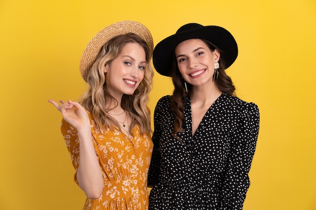 deux jeunes belles femmes amis ensemble isolés sur jaune en robe noire et jaune et chapeau boho élégant s'amusant