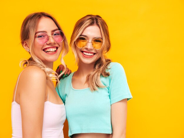 Deux jeunes belles femmes hipster blondes souriantes dans des vêtements d'été à la mode Sexy femmes insouciantes posant près du mur jaune en studio Modèles positifs s'amusant Gai et heureux En lunettes de soleil