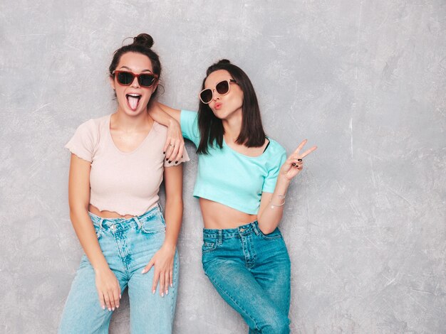 Deux jeunes belles femmes hipster blondes souriantes dans des vêtements d'été à la mode Sexy femmes insouciantes posant près d'un mur gris en studio Modèles positifs s'amusant Gai et heureux en lunettes de soleil