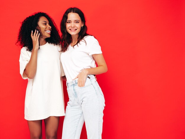 Deux jeunes belles femmes hipster internationales souriantes dans des vêtements d'été à la mode Sexy femmes insouciantes posant près du mur rouge en studio Modèles positifs s'amusant Concept d'amitié