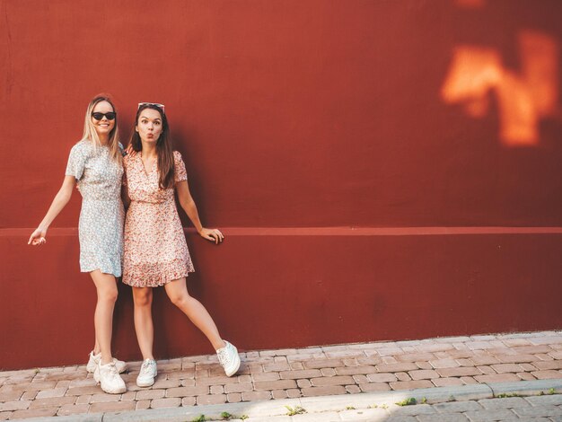 Deux jeunes belles femmes hipster souriantes dans des robes d'été à la modeFemmes insouciantes sexy posant sur le fond de la rue près du mur rouge Modèles purs positifs s'amusant au coucher du soleil étreignant