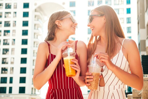 Deux jeunes belles femmes hipster souriantes dans des vêtements d'été à la mode Femmes insouciantes posant à l'extérieurModèles positifs tenant et buvant un smoothie cocktail frais dans une tasse en plastique avec de la paille