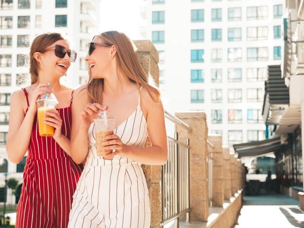 Deux jeunes belles femmes hipster souriantes dans des vêtements d'été à la mode Femmes insouciantes posant à l'extérieurModèles positifs tenant et buvant un smoothie cocktail frais dans une tasse en plastique avec de la paille