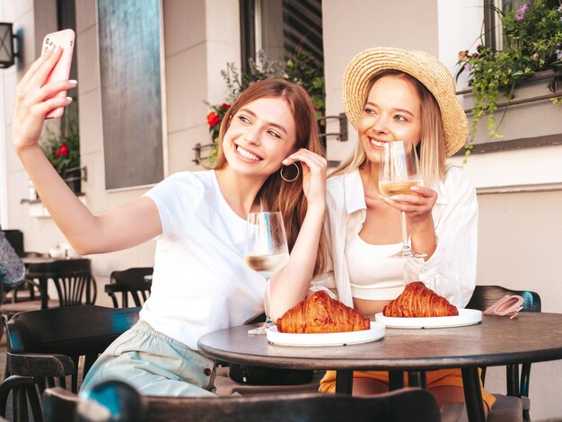 Deux jeunes belles femmes hipster souriantes dans des vêtements d'été à la modeFemmes insouciantes assises au café-véranda dans la rueModèles positifs buvant du vin blancManger un croissant Prendre des photos de selfie