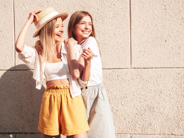 Deux jeunes belles femmes hipster souriantes dans des vêtements d'été à la modeFemmes insouciantes sexy posant dans la rue près d'un mur blanc en chapeau Modèles purs positifs s'amusant au coucher du soleil