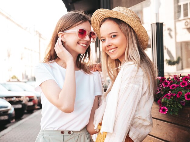 Deux jeunes belles femmes hipster souriantes dans des vêtements d'été à la modeFemmes insouciantes sexy posant sur le fond de la rue en chapeau Modèles purs positifs s'amusant au coucher du soleil étreignant
