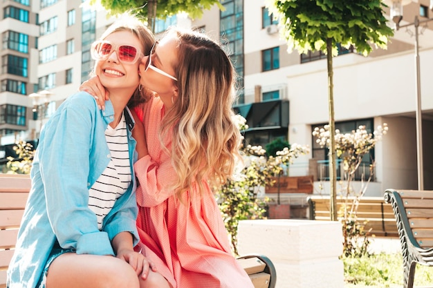 Deux jeunes belles femmes hipster souriantes dans des vêtements et une robe d'été à la modeFemmes insouciantes sexy posant dans la rueModèles purs positifs s'amusantEmbrasser son amie dans la joue