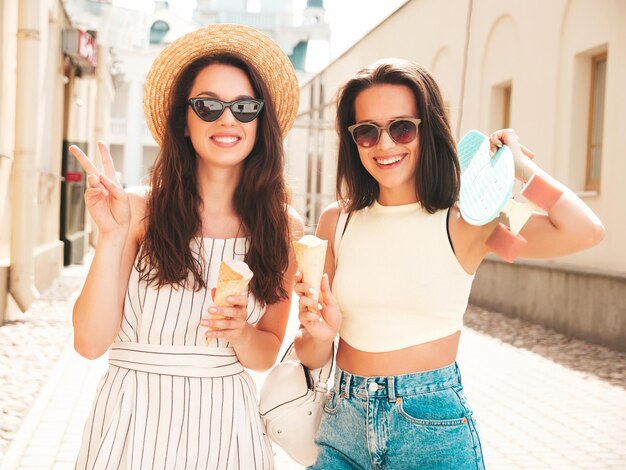 Deux jeunes belles femmes hipster souriantes dans des vêtements et une robe d'été à la modeFemmes insouciantes sexy posant sur le fond de la rue Modèles positifs mangeant de délicieuses glaces dans un cône de gaufres au coucher du soleil