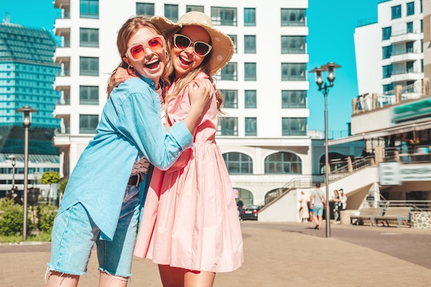 Deux jeunes belles femmes hipster souriantes dans des vêtements et une robe d'été à la modeFemmes insouciantes sexy posant sur le fond de la rue Modèles purs positifs s'amusant au coucher du soleil étreignant et devenant fous