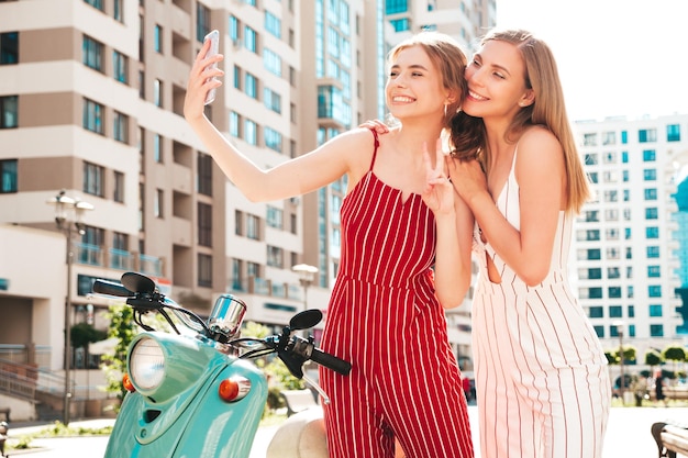 Photo gratuite deux jeunes belles femmes hipster souriantes en salopette à la modefemmes insouciantes sexy conduisant une moto rétro dans la ruemodèles positifs s'amusant sur un scooter italien classiqueprendre des photos de selfie