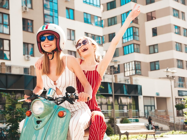 Deux jeunes belles femmes hipster souriantes en salopette à la modeFemmes insouciantes sexy conduisant une moto rétro sur le fond de la rueModèles positifs s'amusant à conduire un scooter italien classique en lunettes