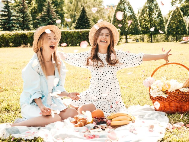 Deux jeunes belles femmes souriantes en robe d'été à la mode et chapeaux. Femmes insouciantes faisant un pique-nique à l'extérieur.