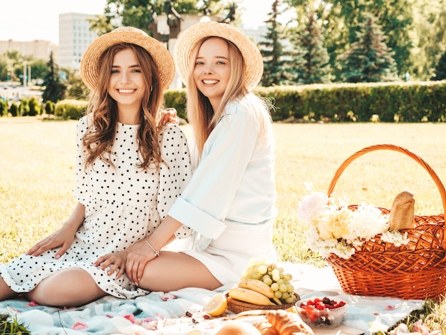 Deux jeunes belles femmes souriantes en robe d'été à la mode et chapeaux. Femmes insouciantes faisant un pique-nique à l'extérieur.