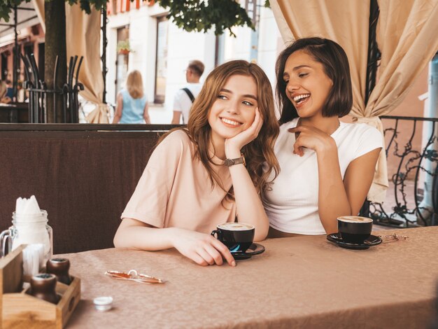 Deux jeunes belles filles hipster souriantes dans des vêtements décontractés d'été à la mode.Des femmes sans soucis discutant dans la terrasse d'un café terrasse et buvant du café.Des modèles positifs s'amusent et communiquent