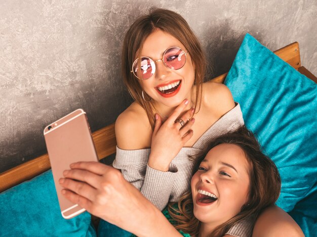 Deux jeunes belles filles magnifiques souriantes dans des vêtements d'été à la mode. Femmes insouciantes sexy posant à l'intérieur et prenant selfie. Modèles positifs s'amusant avec smartphone.