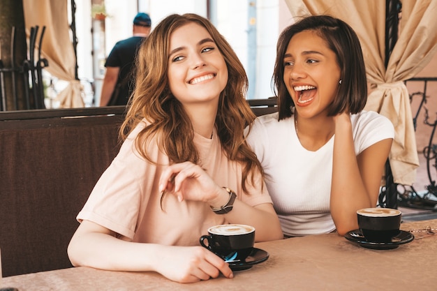 Deux jeunes belles filles souriantes hipster dans des vêtements d'été à la mode.Des femmes sans soucis discutant dans la terrasse d'un café terrasse et buvant du café.Des modèles positifs s'amusent et communiquent