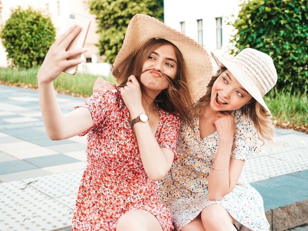 Deux jeunes belles filles souriantes hipster en robe d'été à la mode. Femmes insouciantes sexy assis sur fond de rue en chapeaux. Modèles positifs prenant des photos d'autoportrait selfie sur smartphone