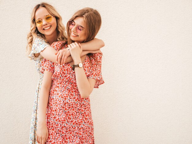 Deux jeunes belles filles souriantes hipster en robe d'été à la mode.Des femmes insouciantes sexy posant dans la rue près du mur à lunettes de soleil. Des mannequins positifs s'amusent et deviennent fous