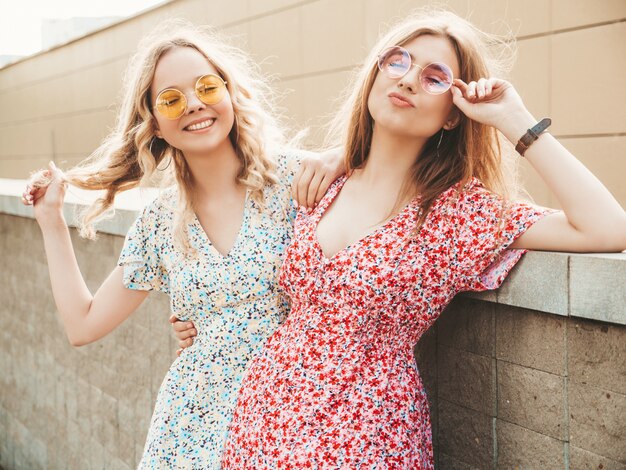 Deux jeunes belles filles souriantes hipster en robe d'été à la mode. Femmes insouciantes sexy posant sur fond de rue en lunettes de soleil. Modèles positifs s'amusant et étreignant