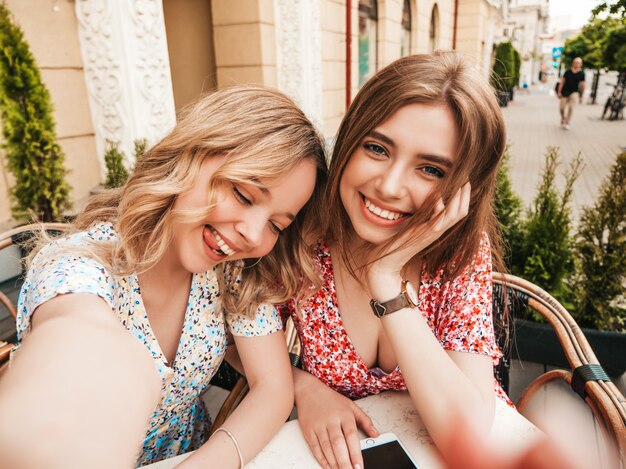Deux jeunes belles filles souriantes hipster en robe d'été à la mode.Des femmes sans soucis discutant dans un café véranda sur le fond de la rue.Des modèles positifs s'amusant et prenant selfie sur smartphone