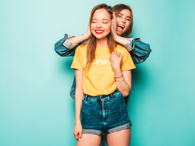 Deux jeunes belles filles souriantes de hipster en T-shirts jaunes d'été et veste en jean. Femmes insouciantes sexy posant près du mur bleu. Modèles branchés et positifs s'amusant