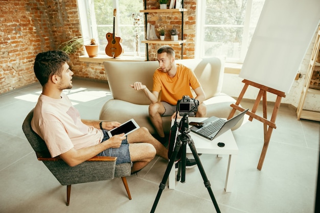 Deux jeunes blogueurs masculins de race blanche dans des vêtements décontractés avec un équipement professionnel ou une caméra d'enregistrement vidéo interview à la maison. Blog, vidéoblog, vlog. Parler en streaming en direct à l'intérieur.