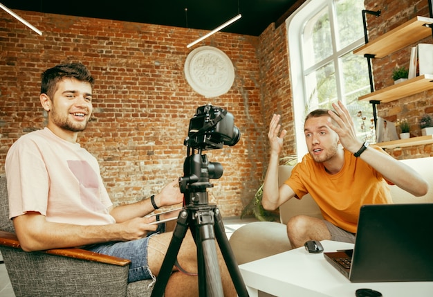 Photo gratuite deux jeunes blogueurs masculins de race blanche dans des vêtements décontractés avec un équipement professionnel ou une caméra d'enregistrement vidéo interview à la maison